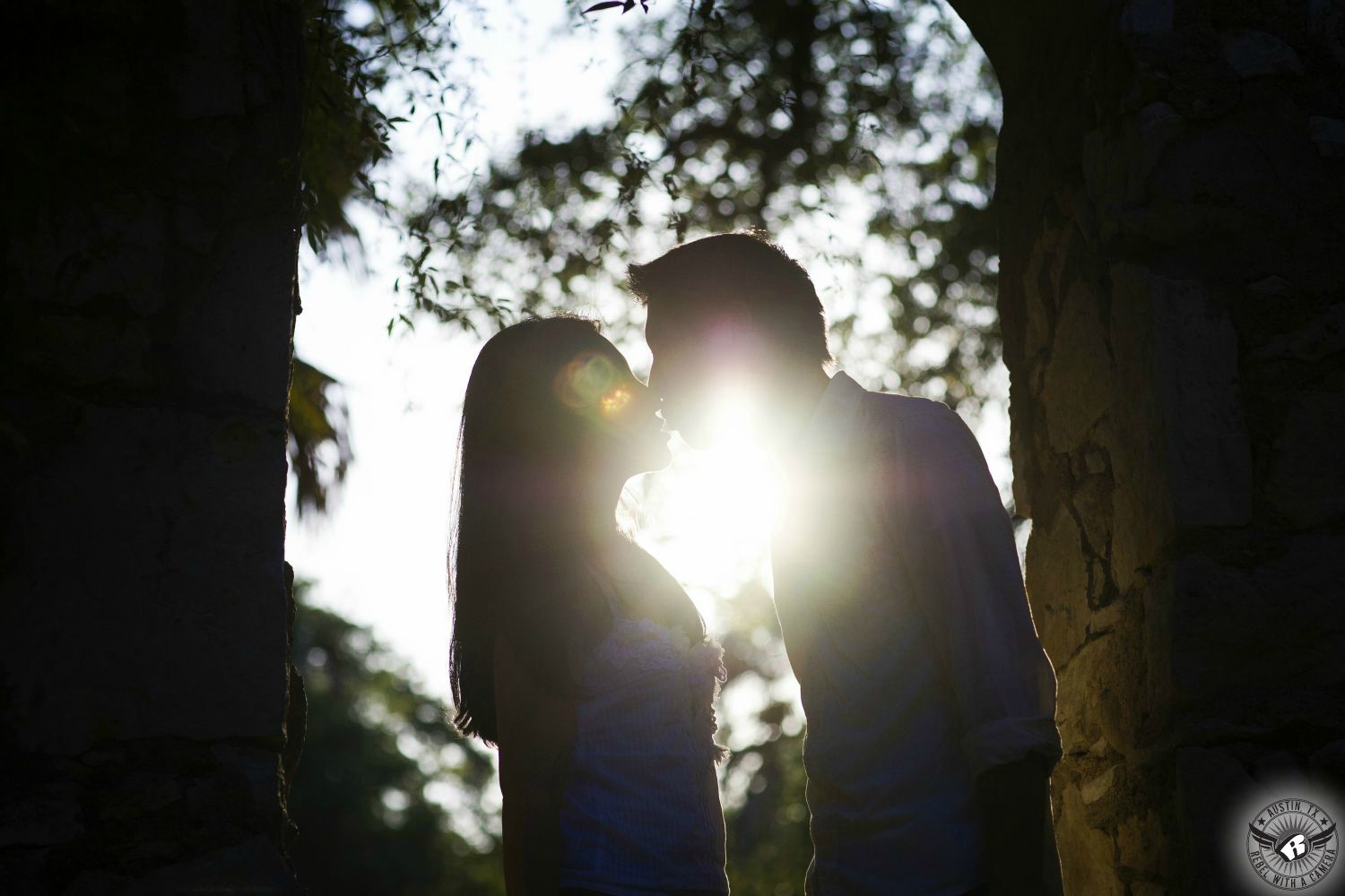 Silhouette of engagement couple kissing with the sun and lens flare in a stone arch with blue sky and blurry trees in the background at Mayfield Park in this tender engagement photograph in the City of Austin.  